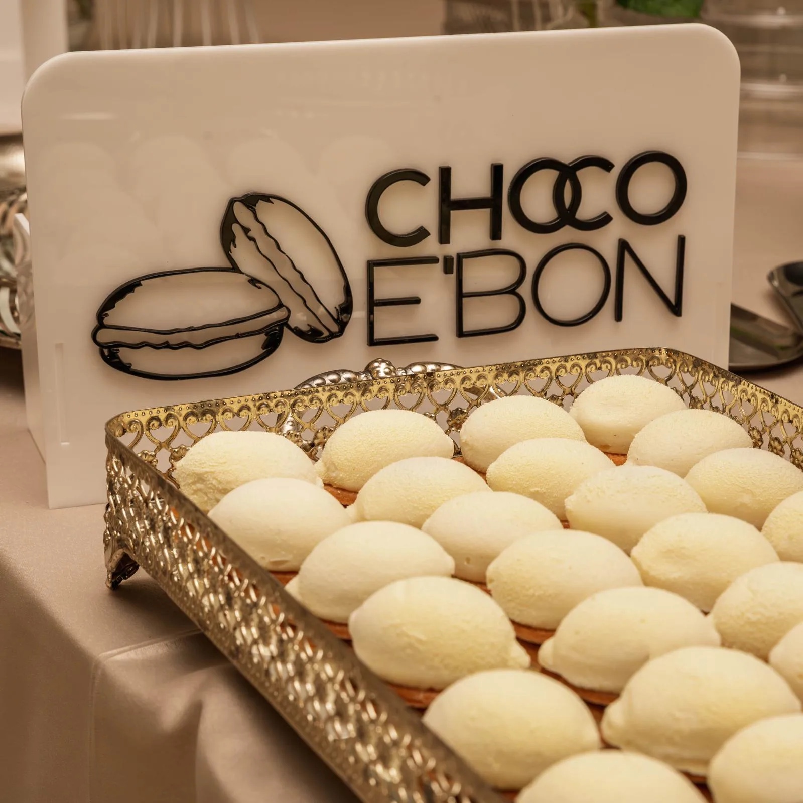Prăjituri albe pe o tavă aurie, în fața plăcuței Choco e'Bon